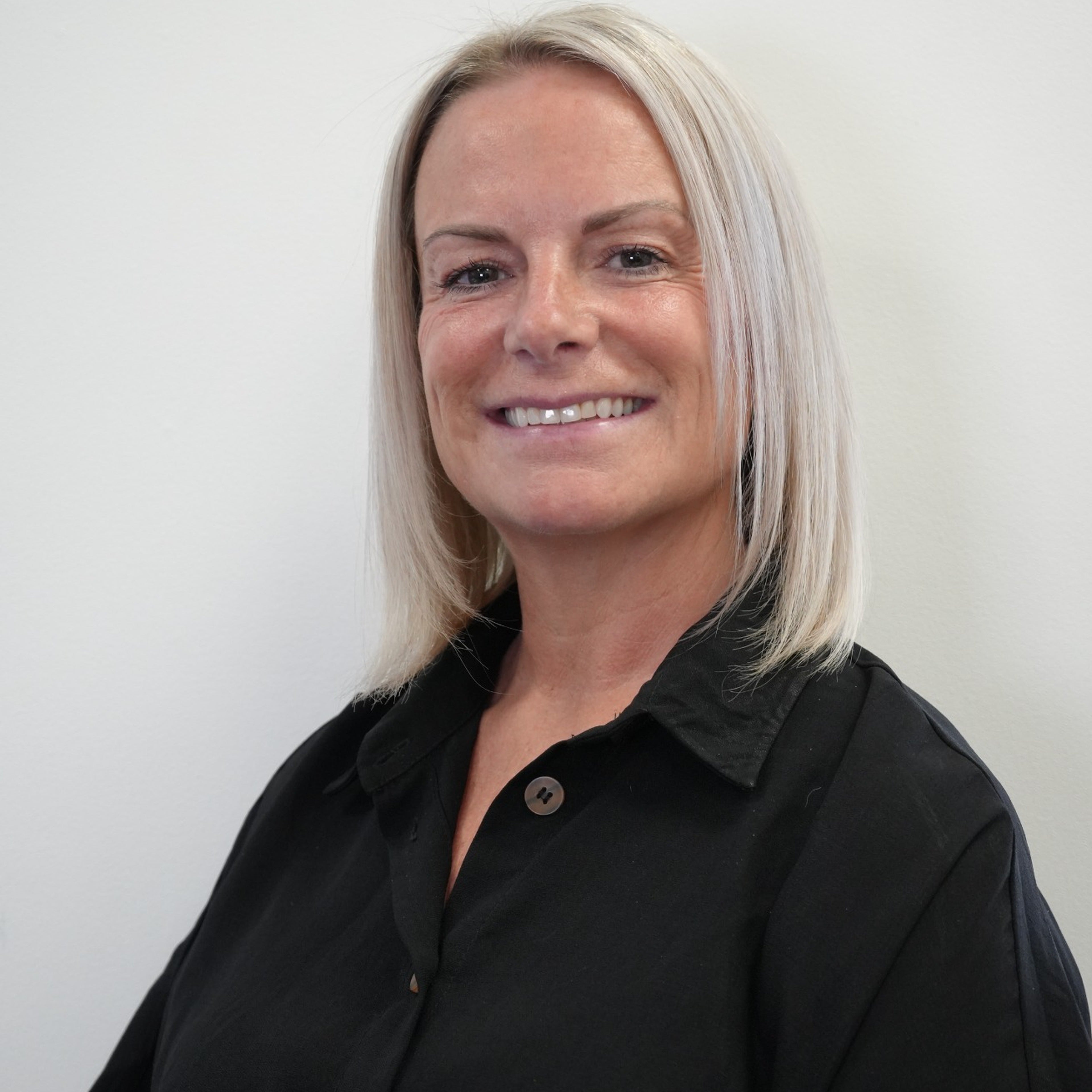 A smiling blonde woman wearing a black short-sleeve button-up shirt, photographed against a plain light gray wall.